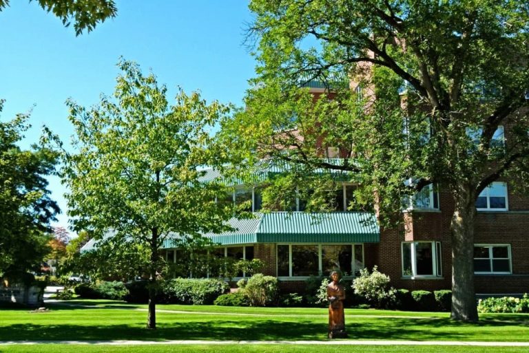 Exterior of brick retirement residence building in Orangeville with green awnings, mature shade trees, landscaped lawn, and accessible entrance for senior community living