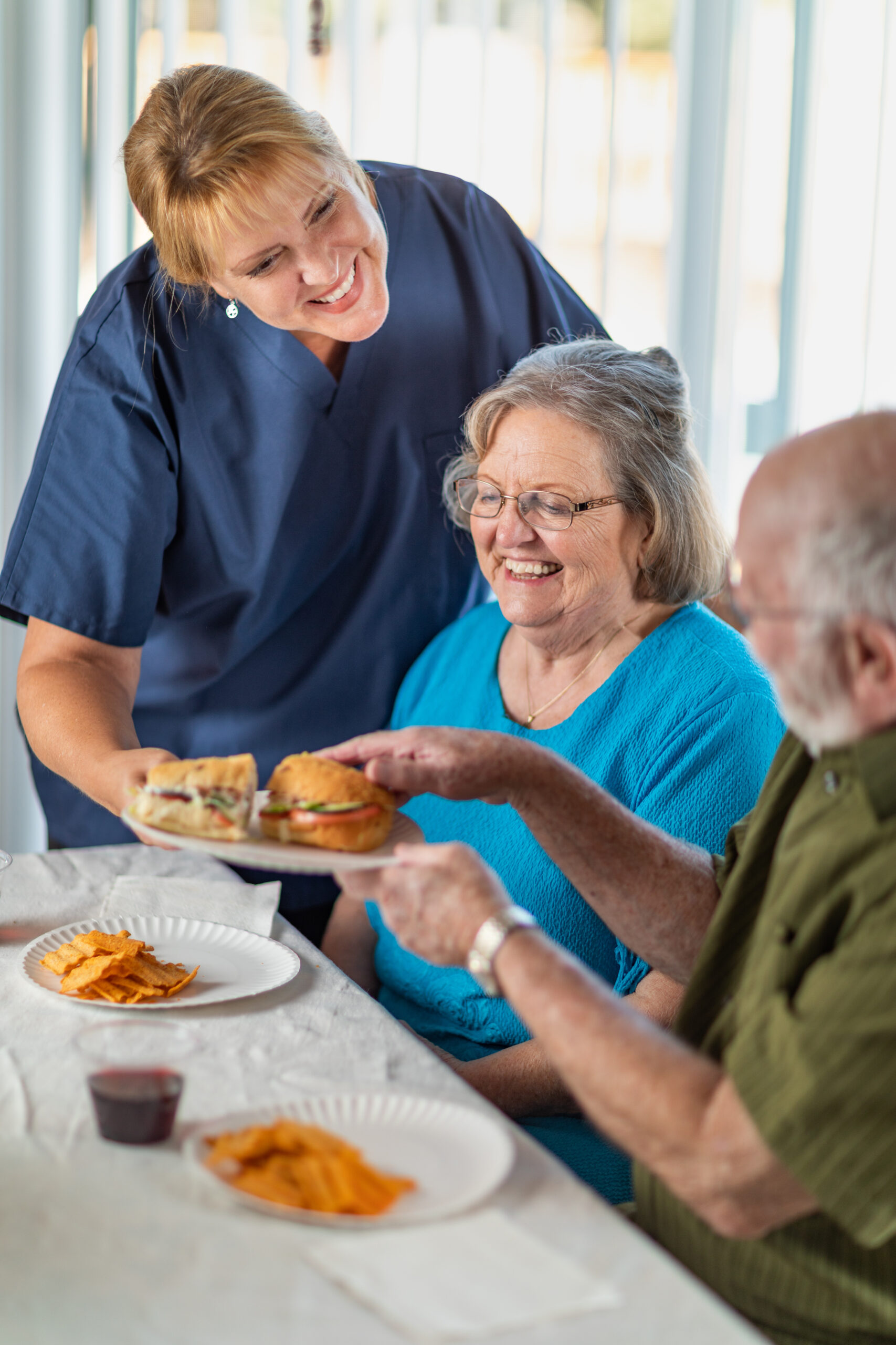 Female Doctor or Nurse Serving Senior Adult Couple Sandwiches at Table.