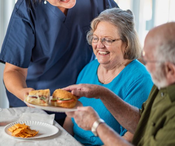 Female Doctor or Nurse Serving Senior Adult Couple Sandwiches at Table.