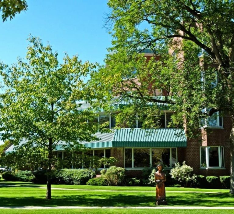 Exterior of brick retirement residence building in Orangeville with green awnings, mature shade trees, landscaped lawn, and accessible entrance for senior community living